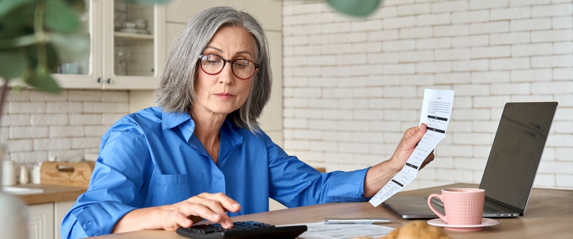 Adult senior 60s woman calculating paper documents at home at pc. Serious middle aged woman at table working on bank payments household online with computer technologies.